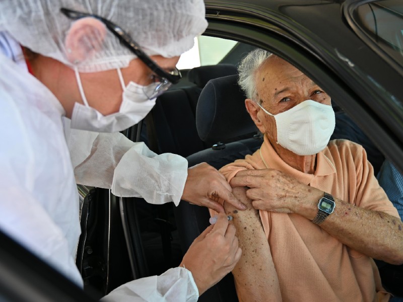 Senhor idoso sentado no banco do passageiro recebe a vacina no braço. Ele está de máscara e camiseta bege. A profissional de saúde usa toca, máscara e avental e está segurando a seringa no braço do vacinado